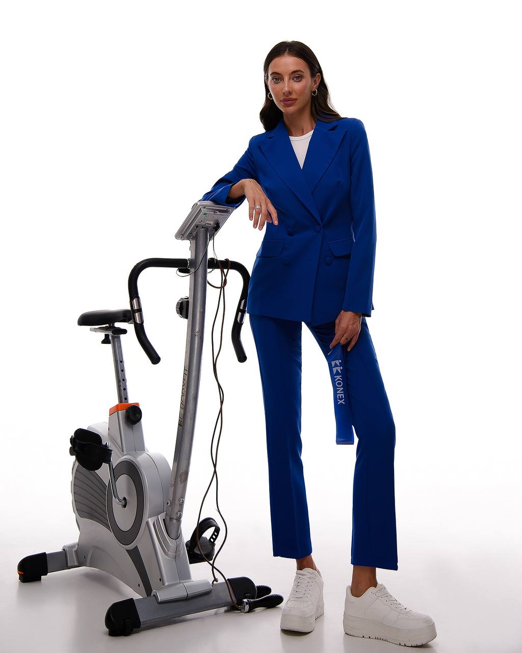Woman in a blue suit standing next to an exercise bike on a white background