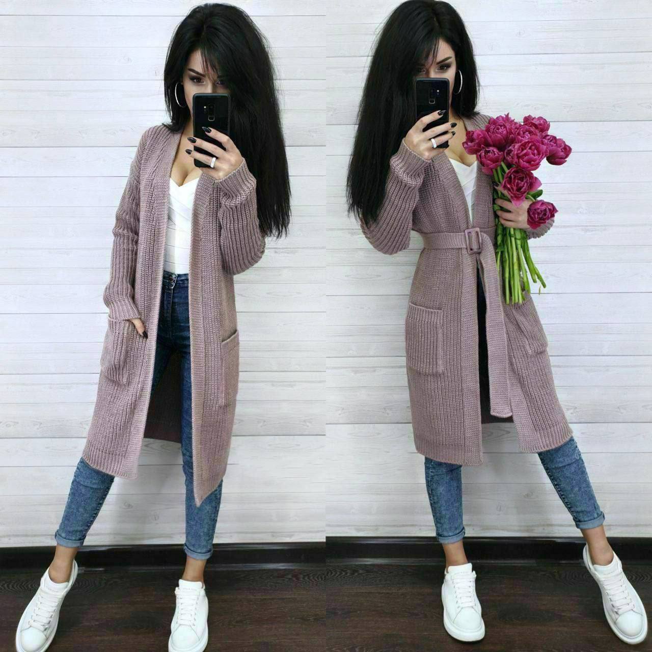 Woman wearing a long pink cardigan holding flowers against a white wall.