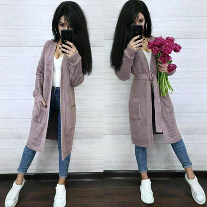 Woman wearing a long pink cardigan holding flowers against a white wall.