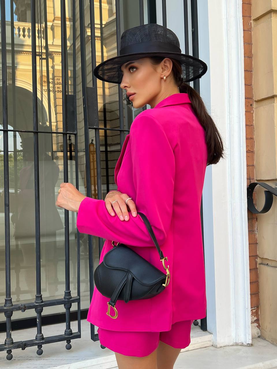 Woman in a bright pink outfit with a black hat and bag, standing in front of a building entrance.