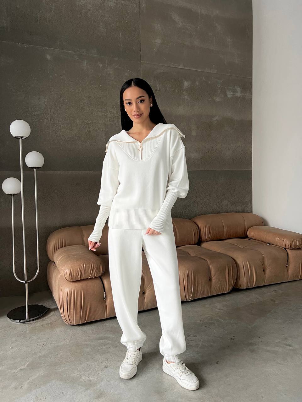Woman in a white outfit standing in a modern living room with a brown sofa and decorative lamp.
