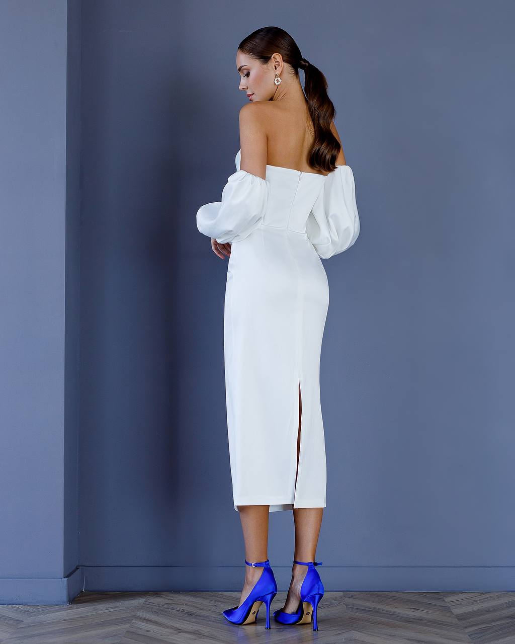 Woman in a white off-shoulder dress with puffed sleeves against a blue wall.