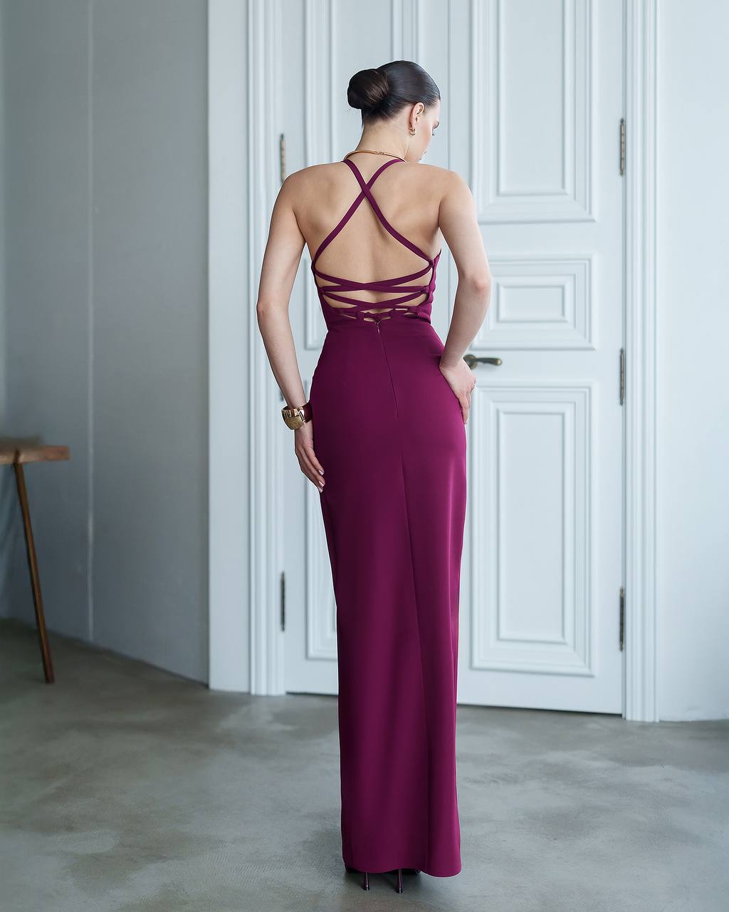 Woman wearing a burgundy evening gown with a backless design in a room with white walls and a door.