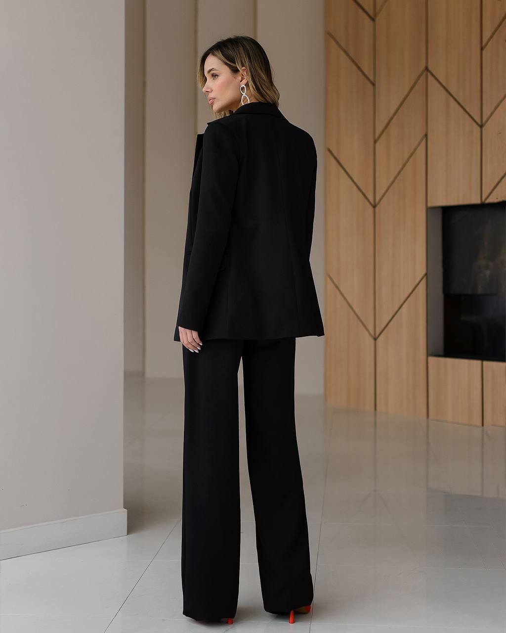 Woman in a black suit standing in a modern interior setting with wooden wall and white floor.