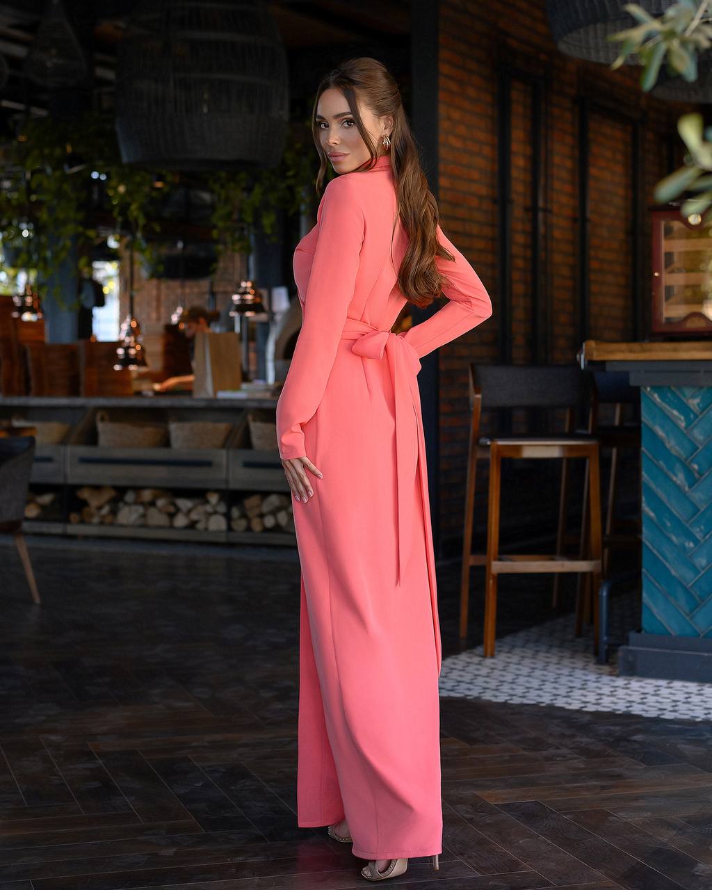 Woman in a pink dress standing in an indoor setting with wooden furniture and plants.