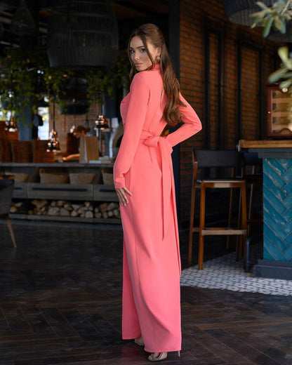 Woman in a pink dress standing in an indoor setting with wooden furniture and plants.