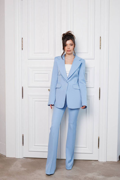 Woman wearing a light blue suit standing against a white paneled wall.