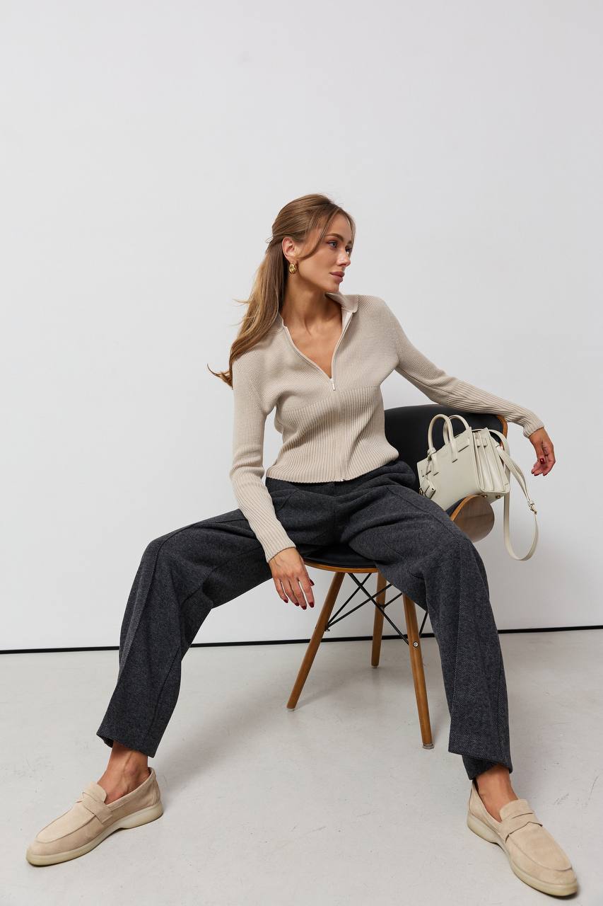 Woman sitting on a chair wearing a beige sweater and dark pants, holding a white handbag.