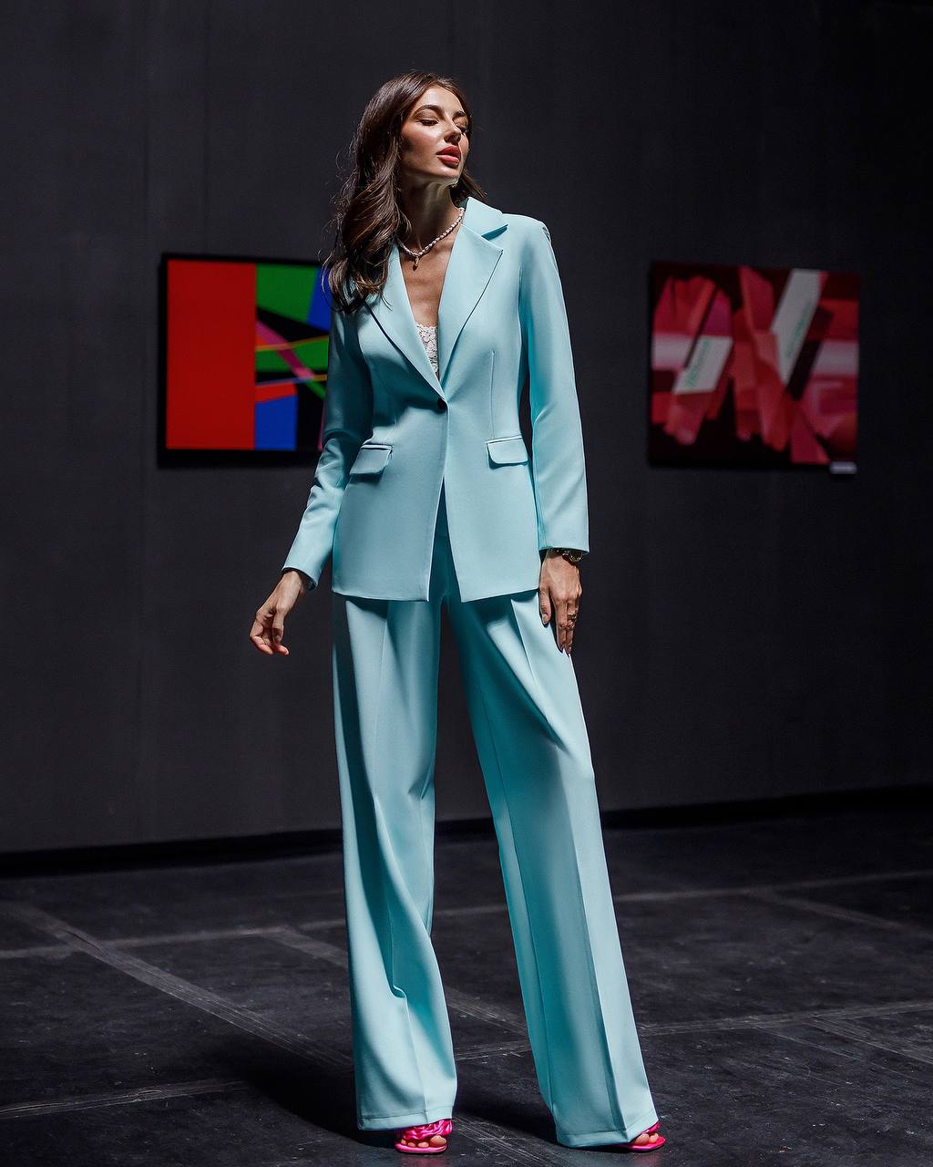 Woman in a light blue suit standing in a dark room with colorful abstract art on the wall.