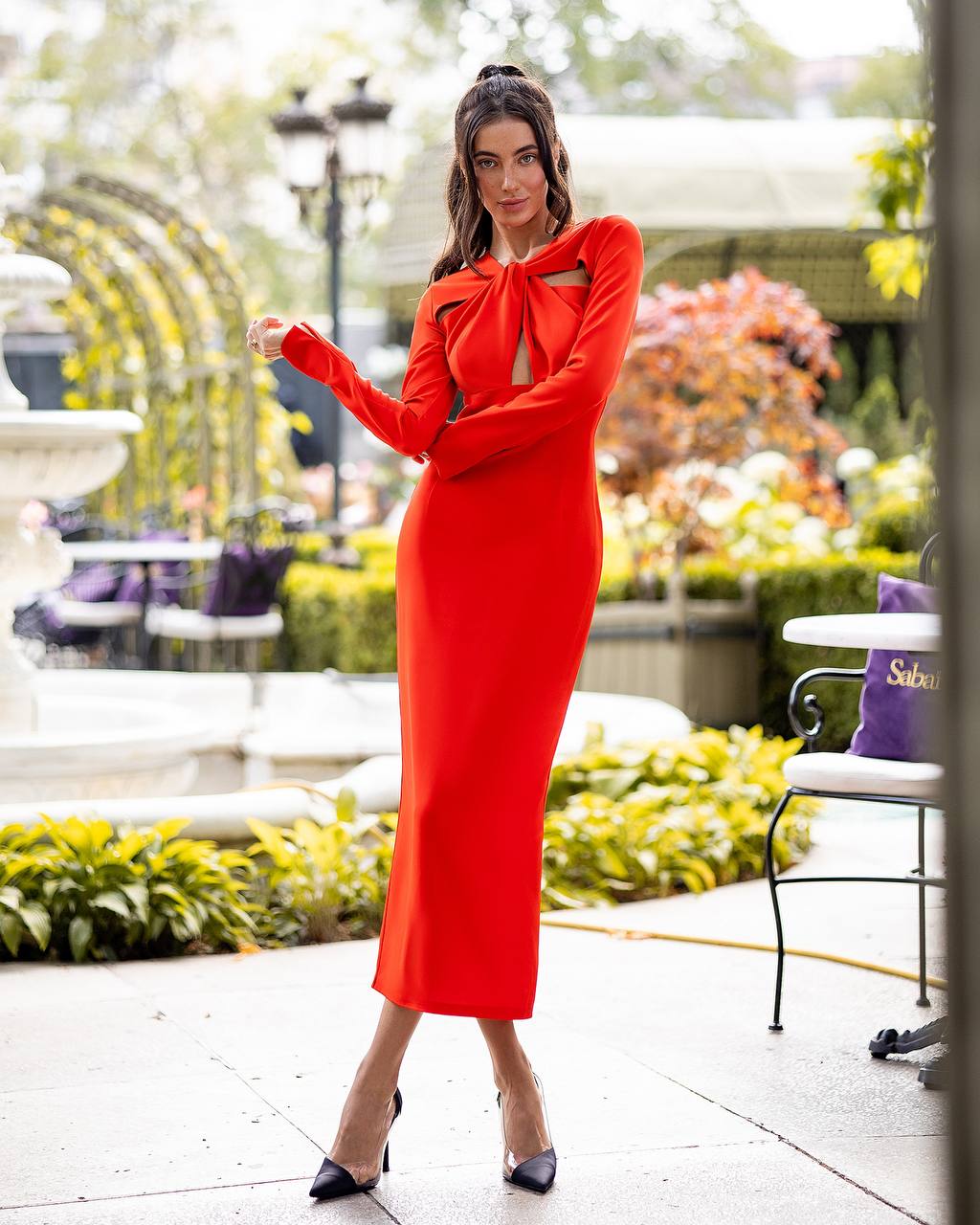 Woman in a red dress standing outdoors with greenery and a building in the background