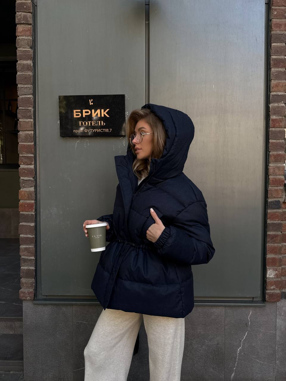 Person wearing a navy puffer jacket holding a coffee cup outside a building with a sign.
