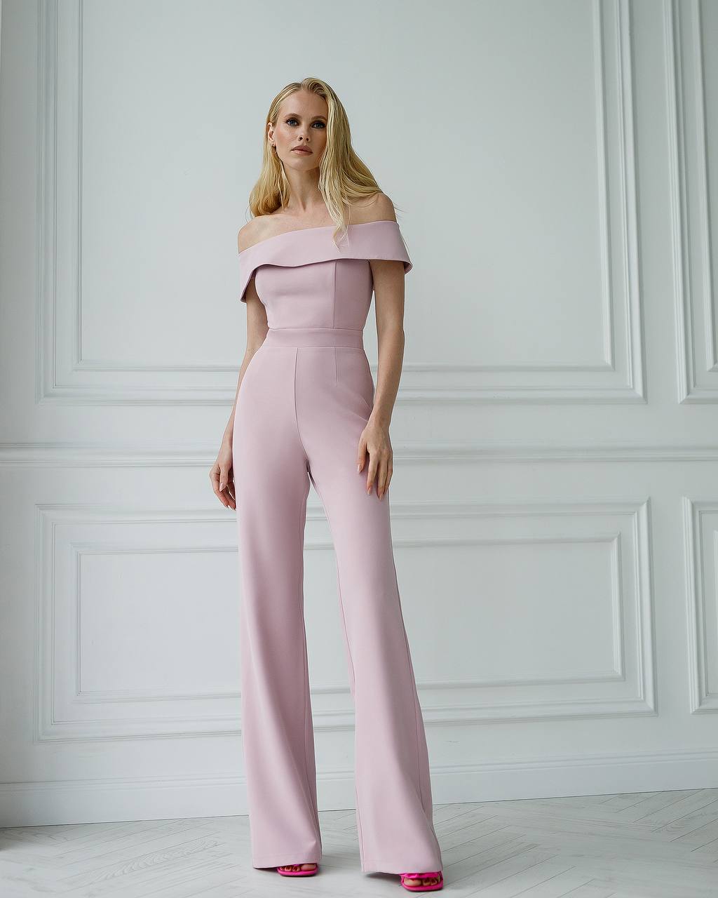 Woman wearing a pink off-shoulder jumpsuit against a white paneled wall.