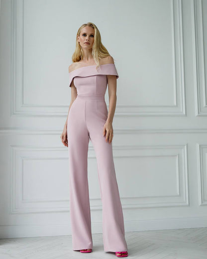 Woman wearing a pink off-shoulder jumpsuit against a white paneled wall.