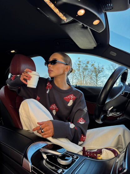 Woman sitting in a car holding a coffee cup, wearing a sweater with mushroom designs.