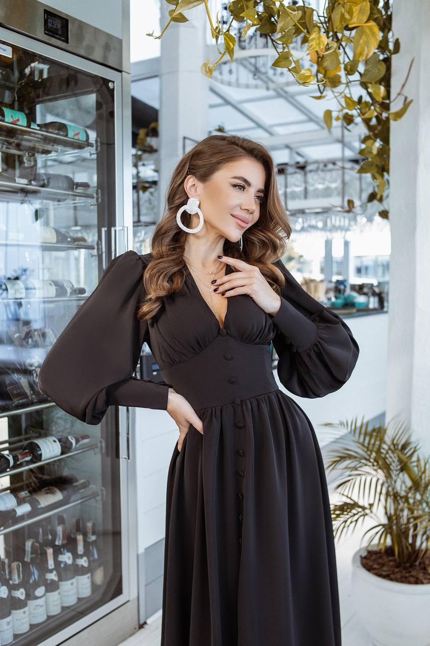 Woman in a black dress standing in a modern kitchen with a refrigerator and plants.
