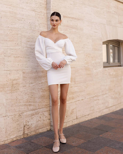 Woman in a white off-shoulder dress standing against a beige stone wall.