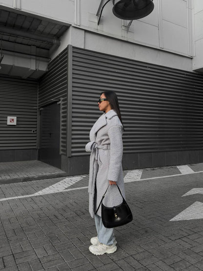 Person wearing a long gray coat and light blue jeans standing on a street.