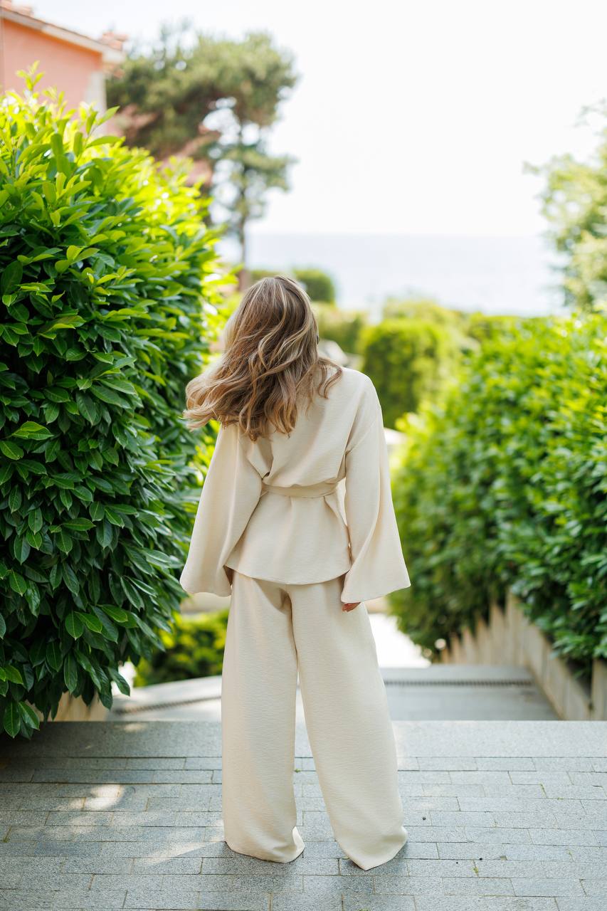 Woman in a beige suit walking away from the camera on a path with greenery.