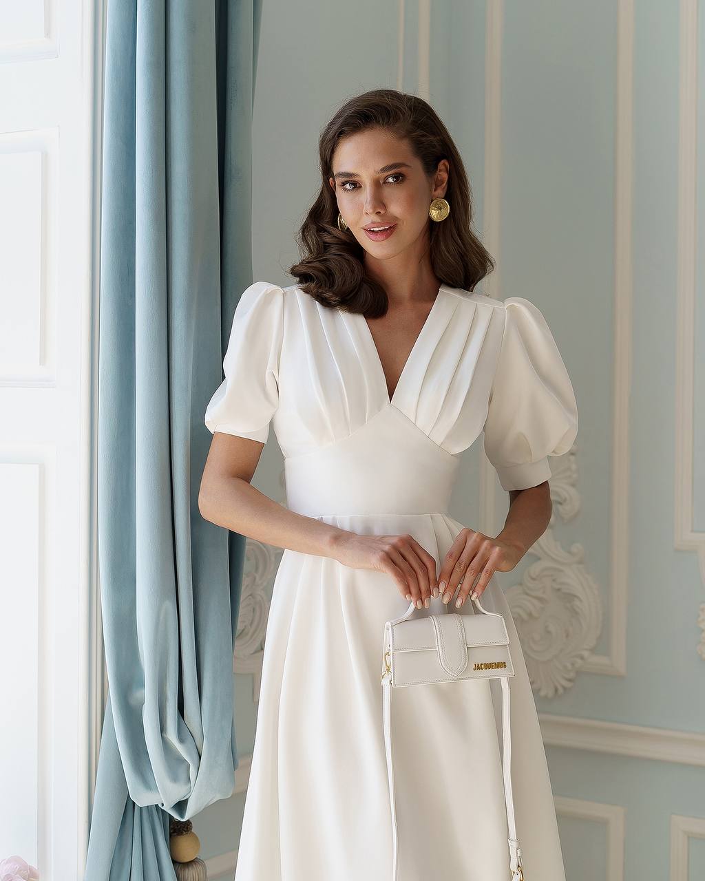 Woman in a white dress holding a white handbag against a decorative wall.