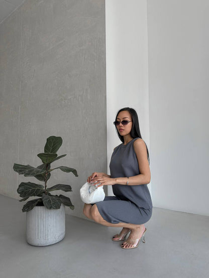 Woman in a gray dress holding a white vase next to a potted plant in a minimalistic setting.