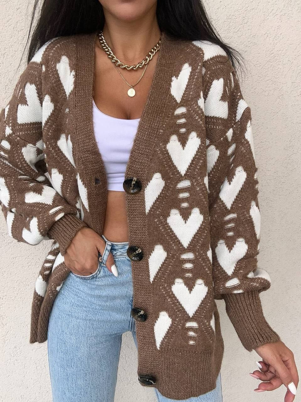 Person wearing a brown cardigan with white heart patterns, white top, and blue jeans against a beige background.