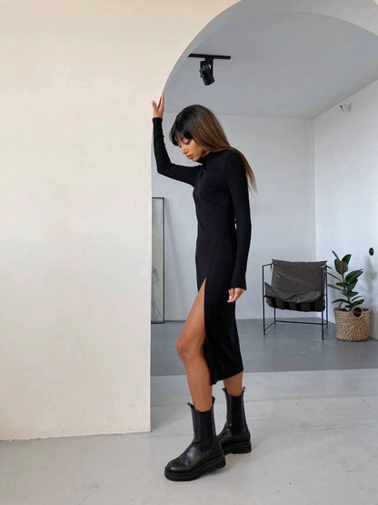Person wearing a black dress with a slit and black boots in a minimalistic room.