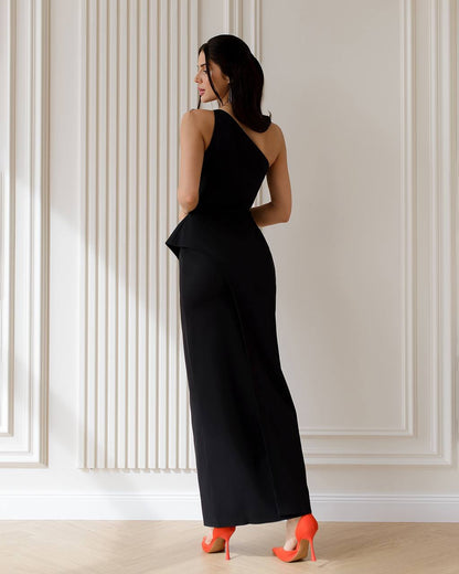 Woman wearing a black evening gown with a high slit, standing against a light-colored paneled wall.