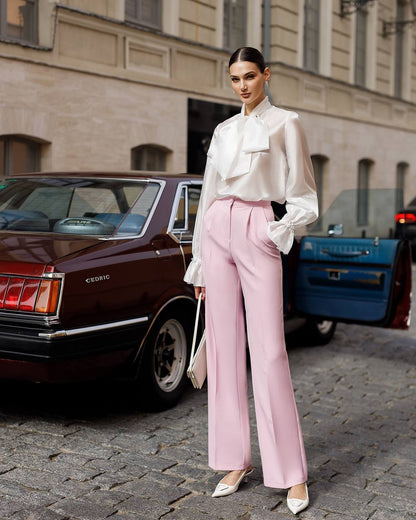 Woman in a white blouse and pink pants standing on a street with vintage cars in the background