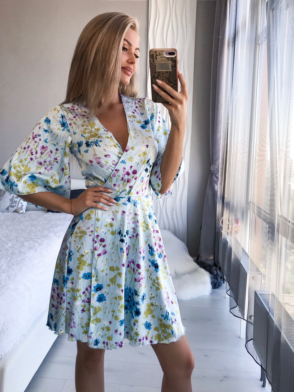 Woman taking a mirror selfie wearing a floral dress in a bedroom setting.