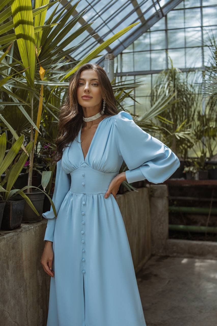 Woman in a light blue dress standing in a greenhouse with plants around.