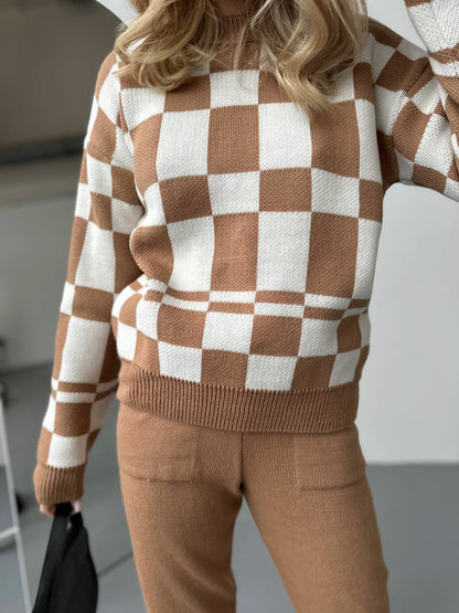 Person wearing a brown and white checkered sweater with beige pants indoors.