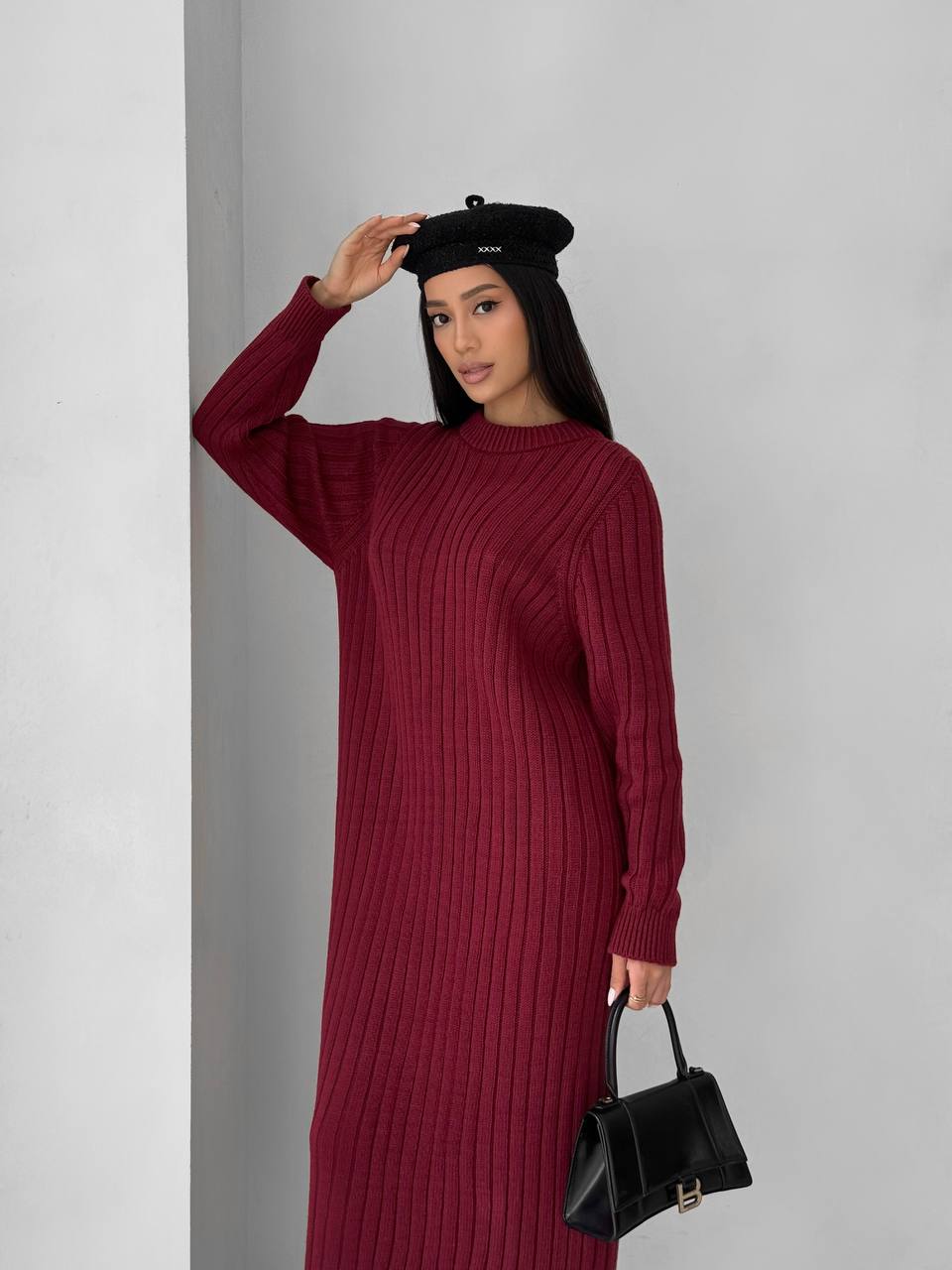 Woman wearing a burgundy ribbed dress and black beret holding a black handbag against a gray background