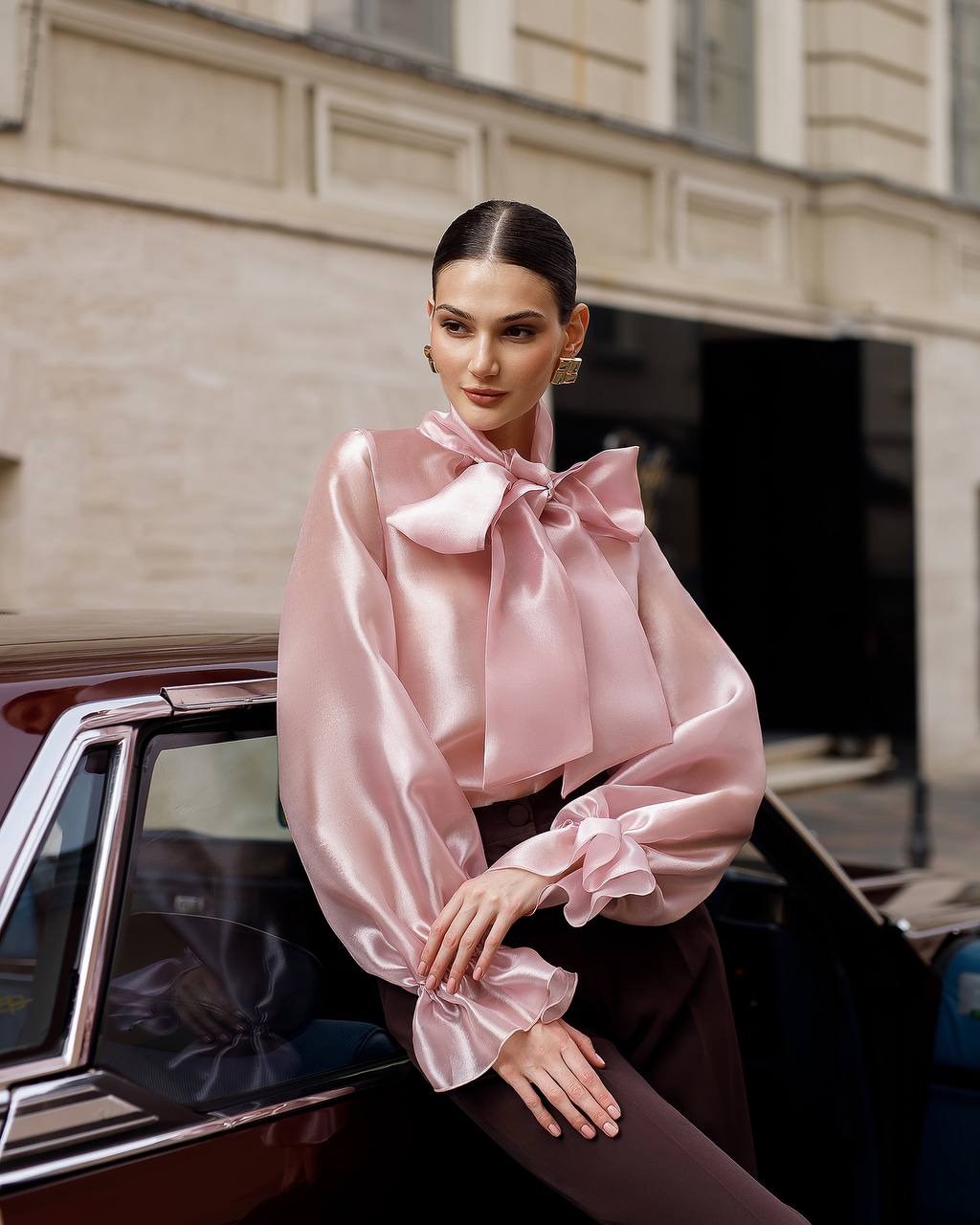 Woman in a pink blouse with a large bow sitting next to a vintage car.