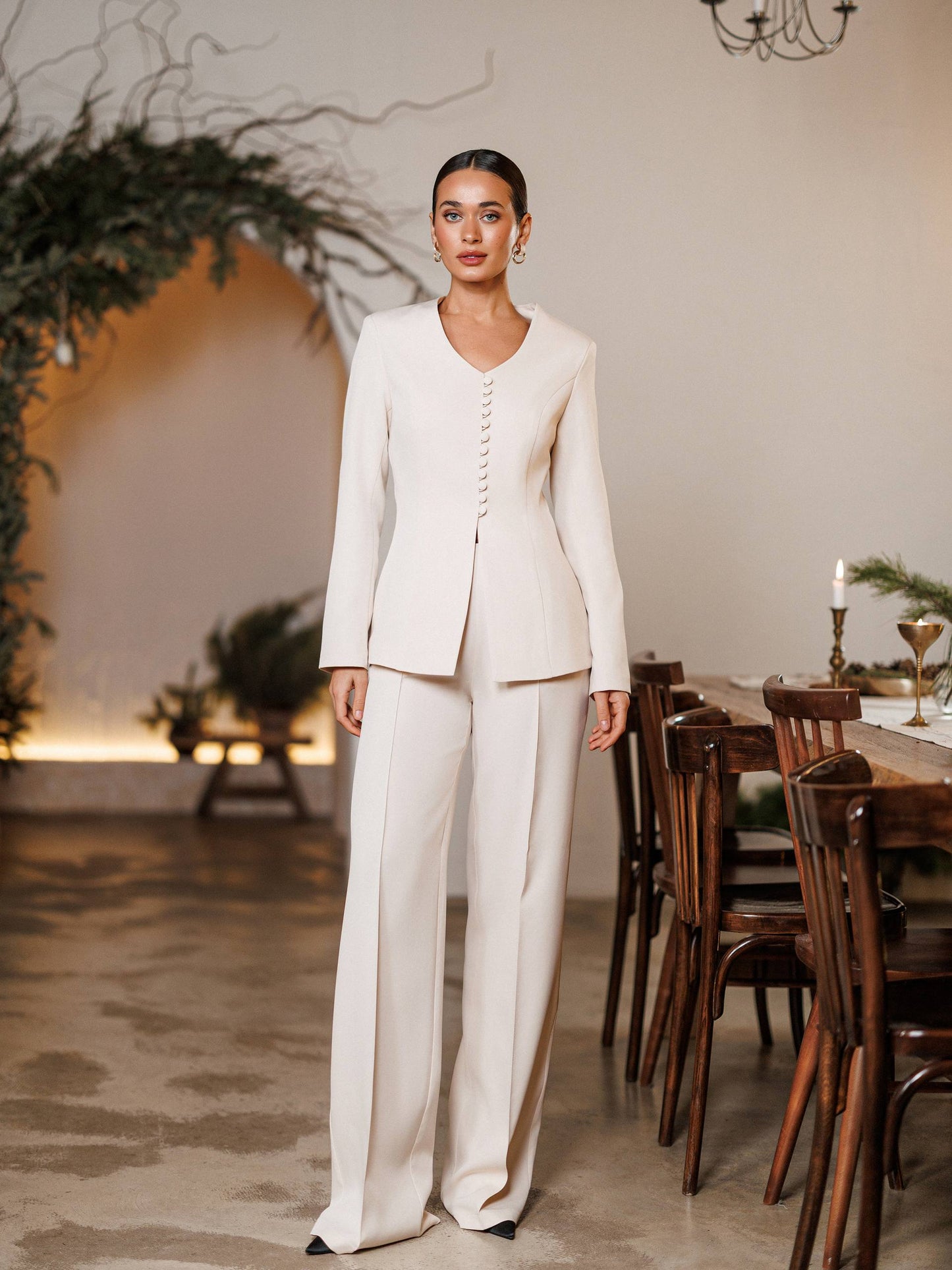 Woman in a white suit standing in a elegant indoor setting with a table and chairs.