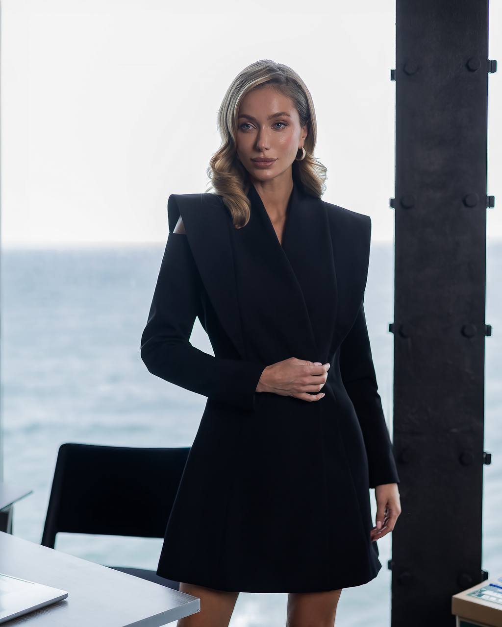 Woman wearing a black coat standing by a window with a view of water.