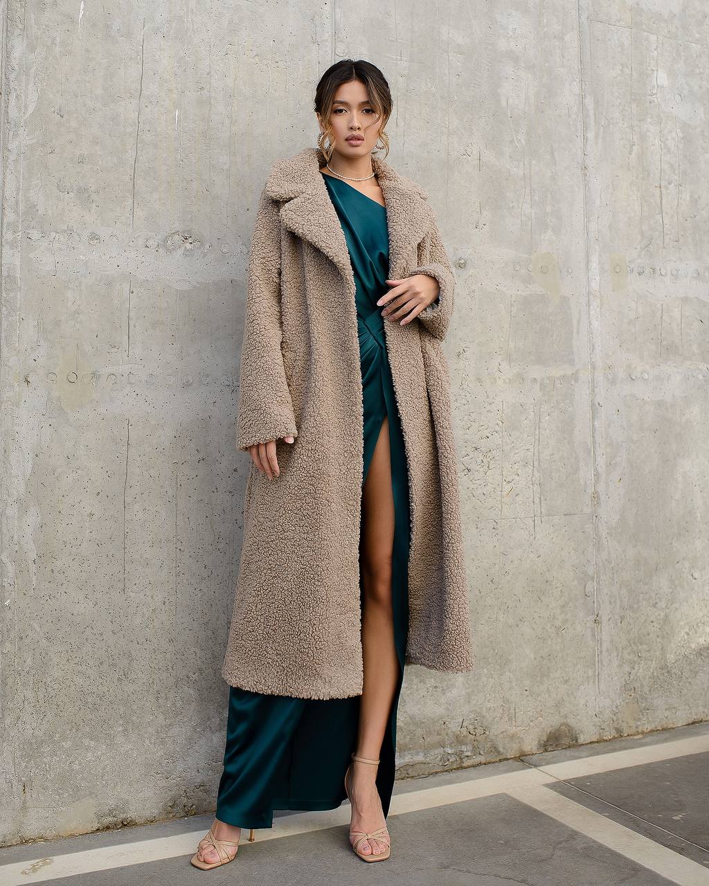 Woman wearing a long beige coat over a teal dress against a concrete wall.