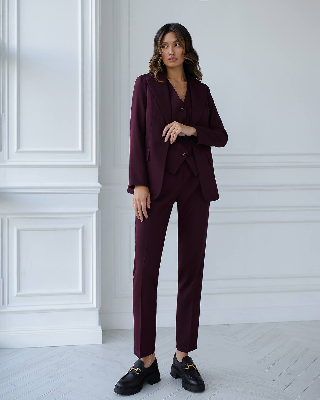 Woman wearing a burgundy suit standing against a white paneled wall.