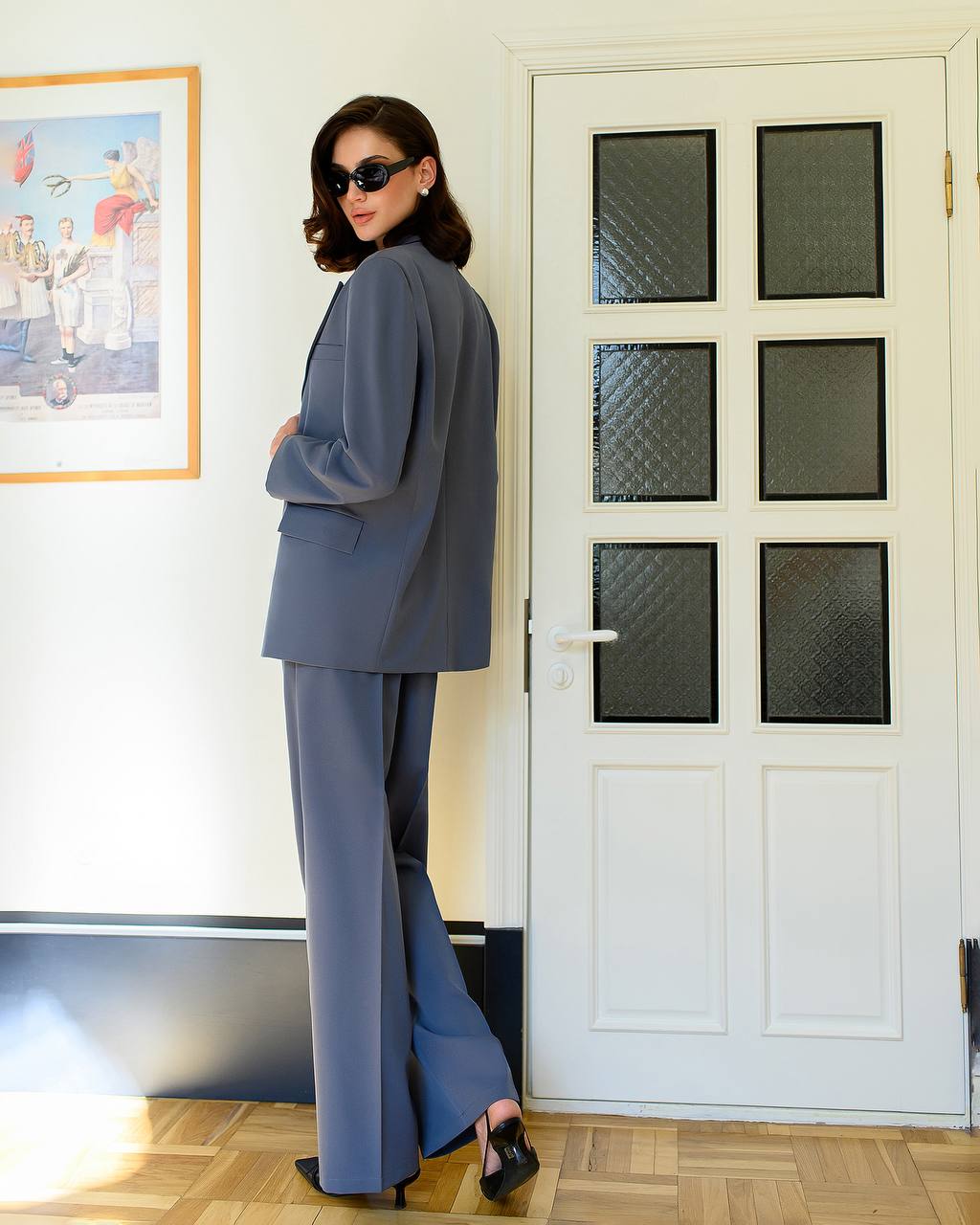 Woman in a gray suit standing in a room with a white door and framed picture on the wall.