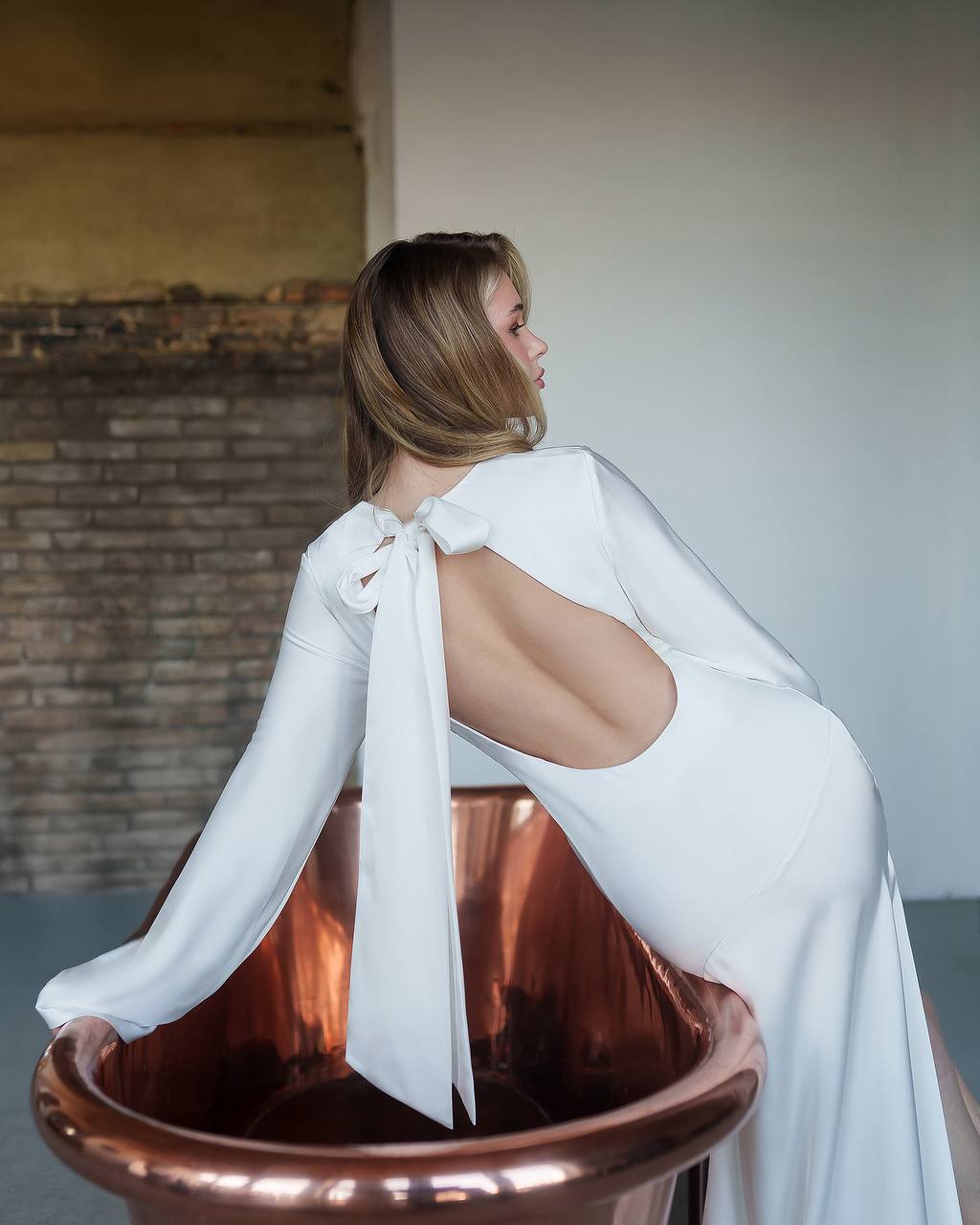 Woman in a white dress sitting in a copper bathtub with a neutral background