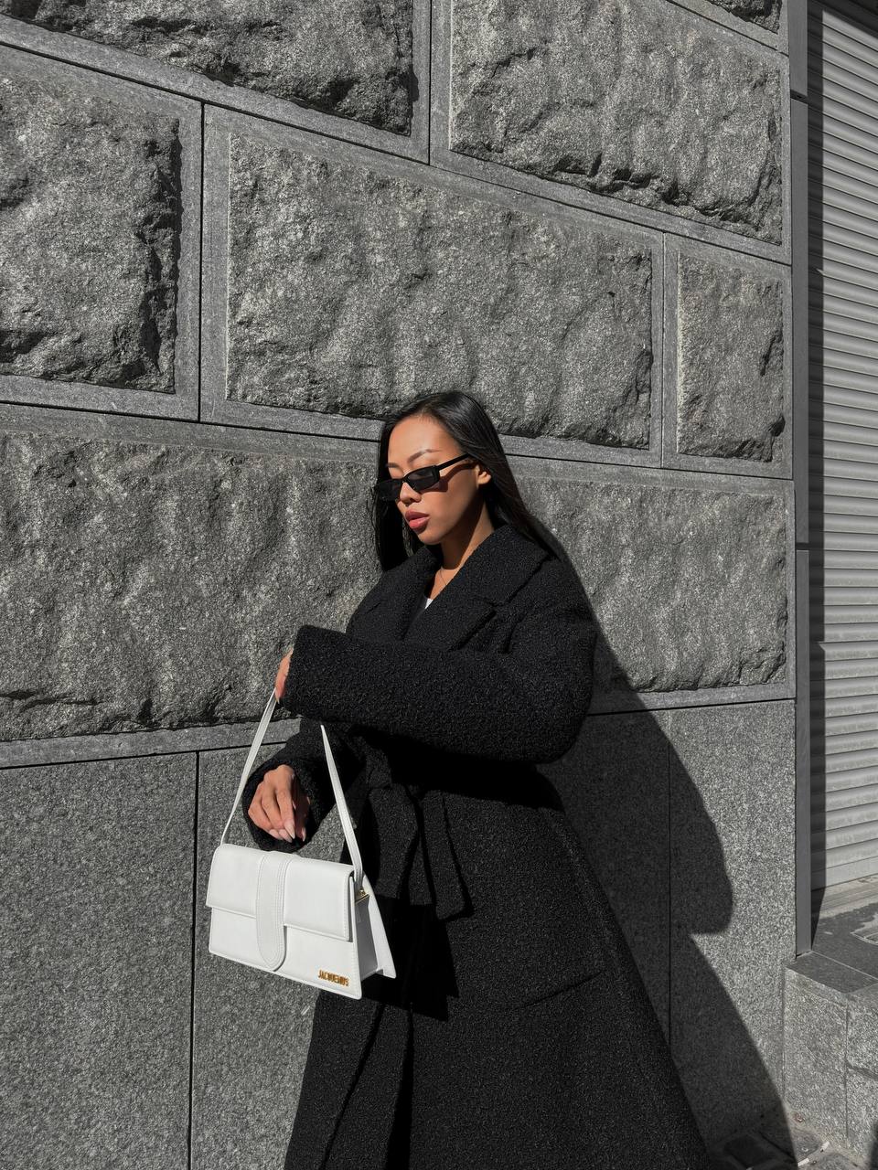 Person in a black coat holding a white bag against a stone wall.