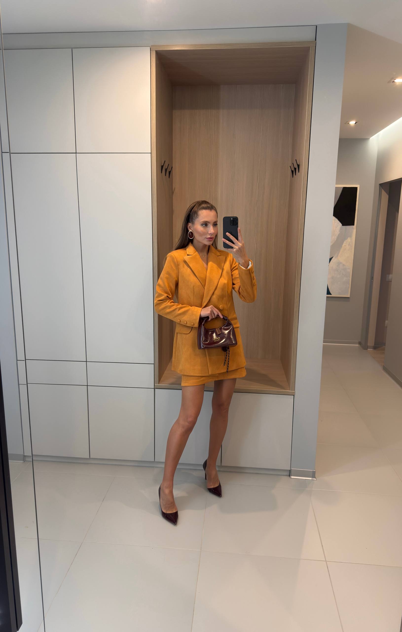 A woman takes a mirror selfie in a modern interior, wearing a mustard yellow blazer and matching mini skirt with dark brown pointed heels and holding a dark brown handbag.