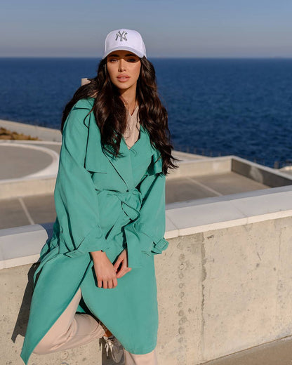 Woman in a teal coat and white cap standing by the ocean.