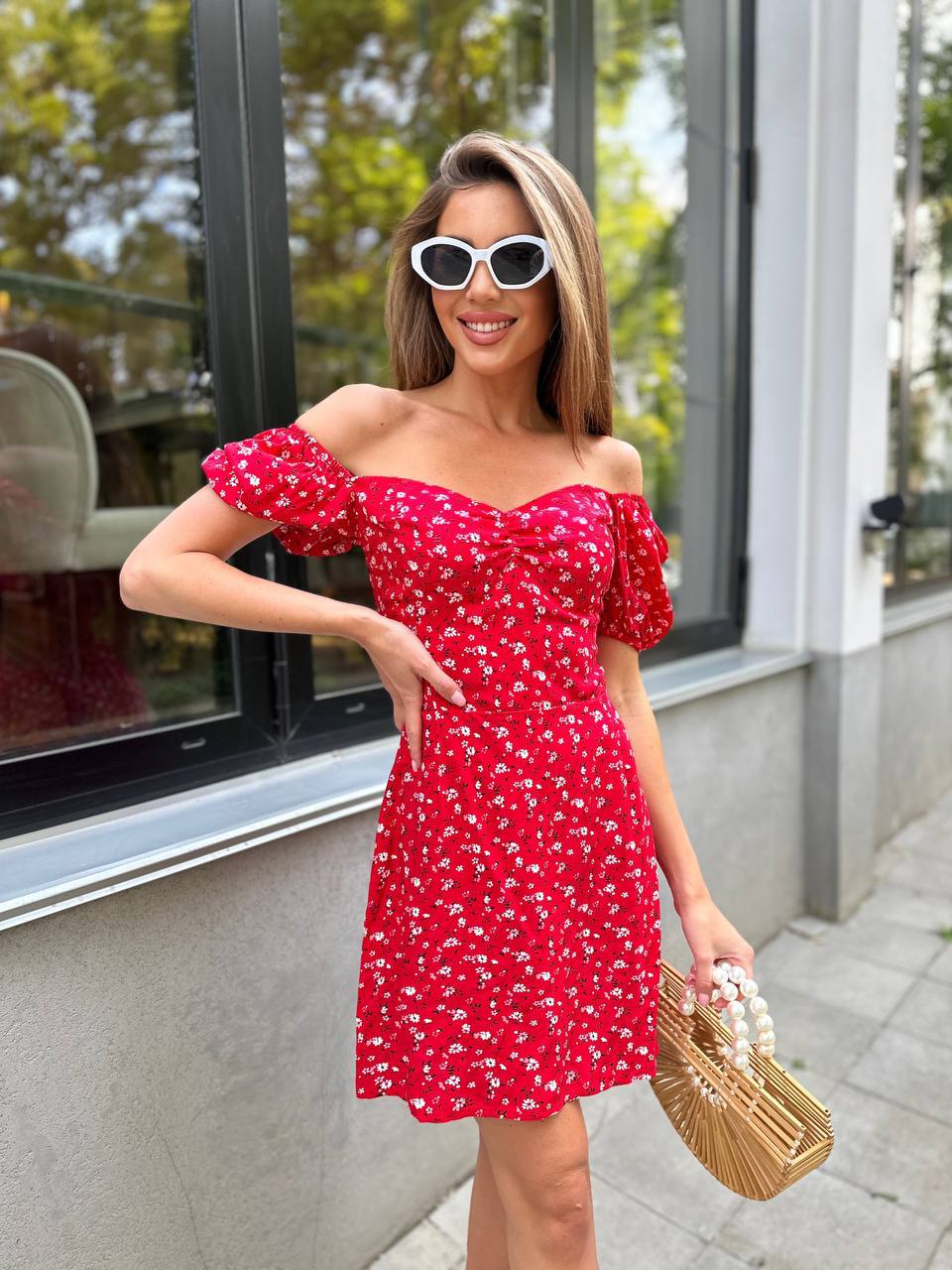 Woman wearing a red floral dress with sunglasses and a straw bag outdoors.