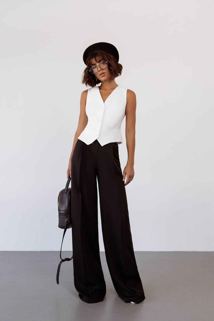 Person wearing a white sleeveless top and black wide-leg pants, holding a black backpack against a white background.