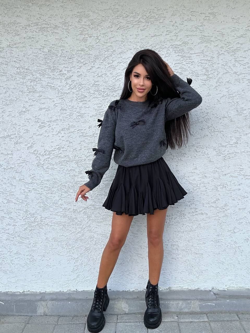Woman wearing a gray sweater and black pleated skirt against a light gray wall.