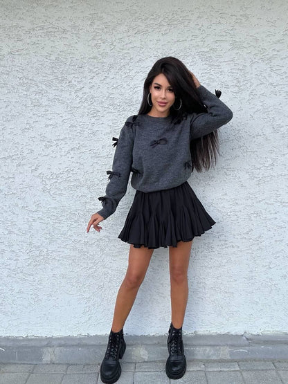 Woman wearing a gray sweater and black pleated skirt against a light gray wall.