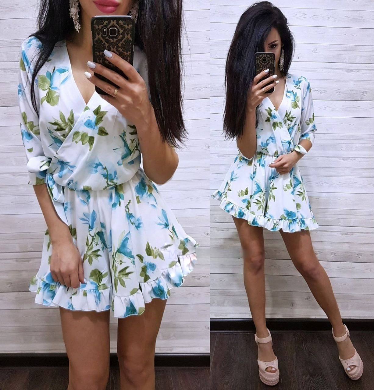 Woman wearing a floral dress taking a mirror selfie against a white wall.