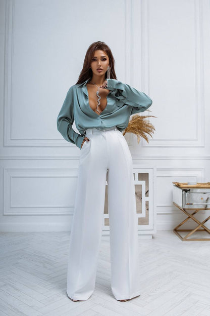 Woman wearing a light blue blouse and white pants in a room with white walls and wooden floor.