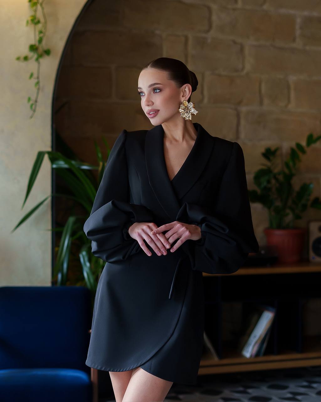 Woman in a black dress standing indoors with plants and a blue couch in the background