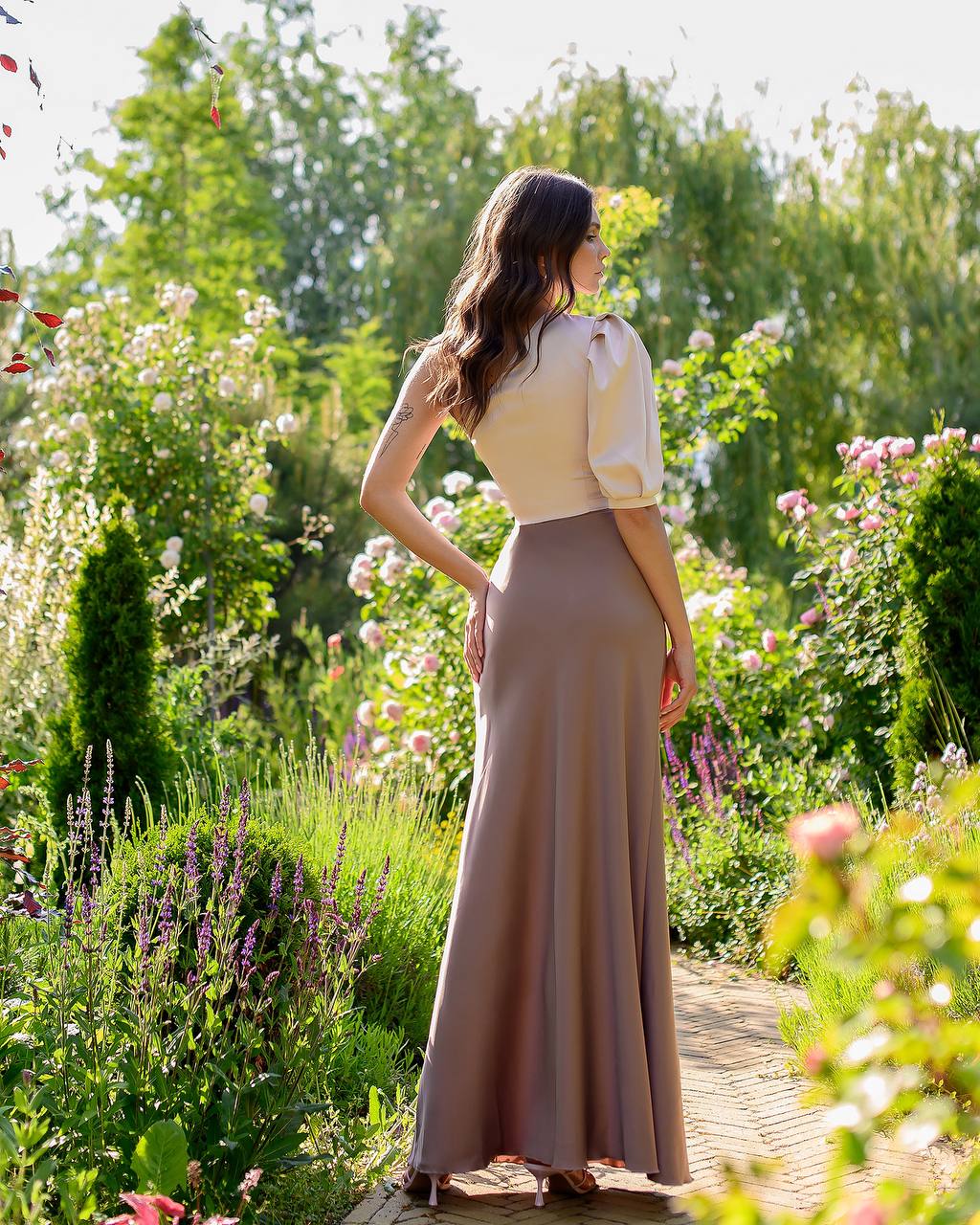 Woman in a long dress standing in a garden with flowers and greenery.